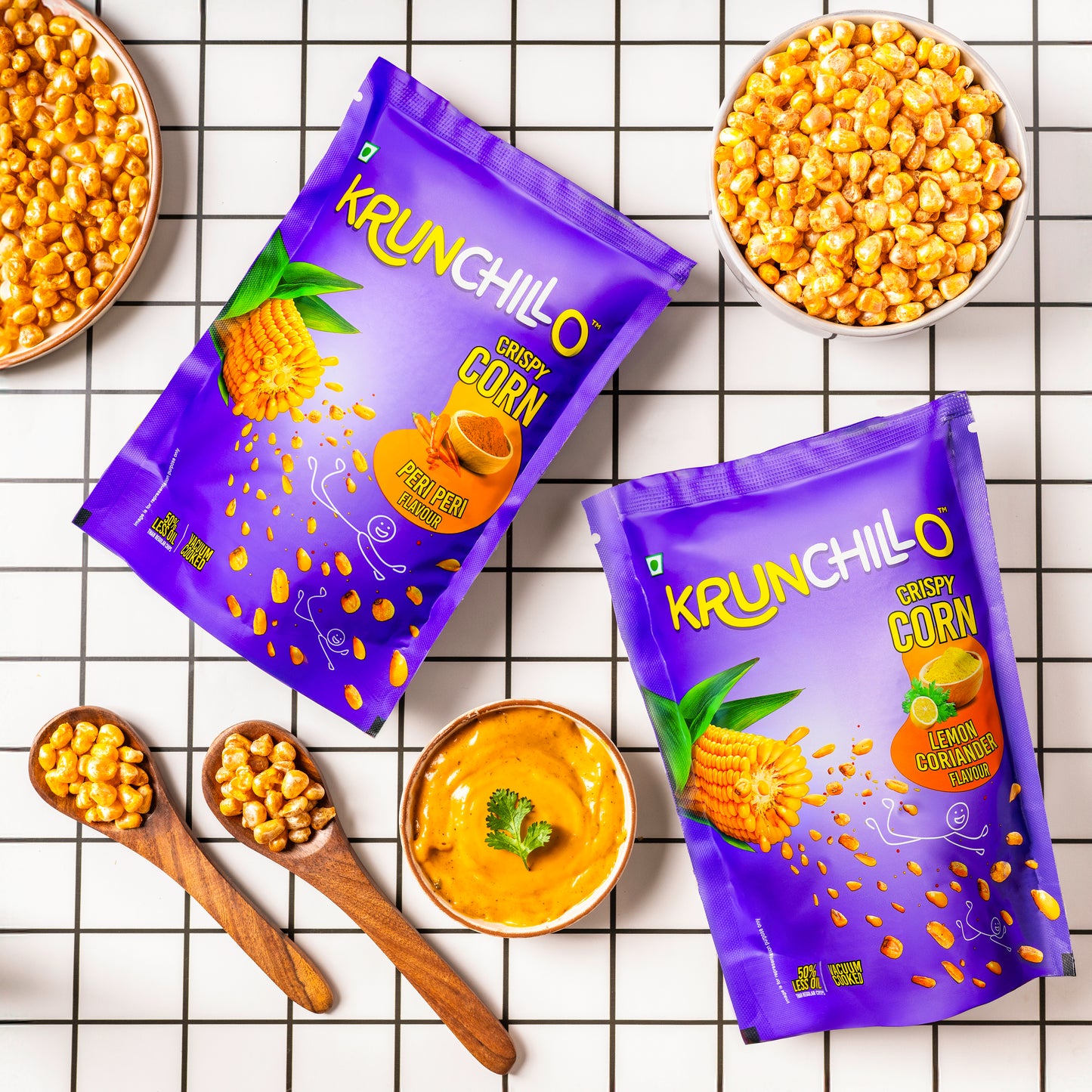 Two purple packages of Krunchillo crispy corn on a checkered background with corn and dipping sauce.